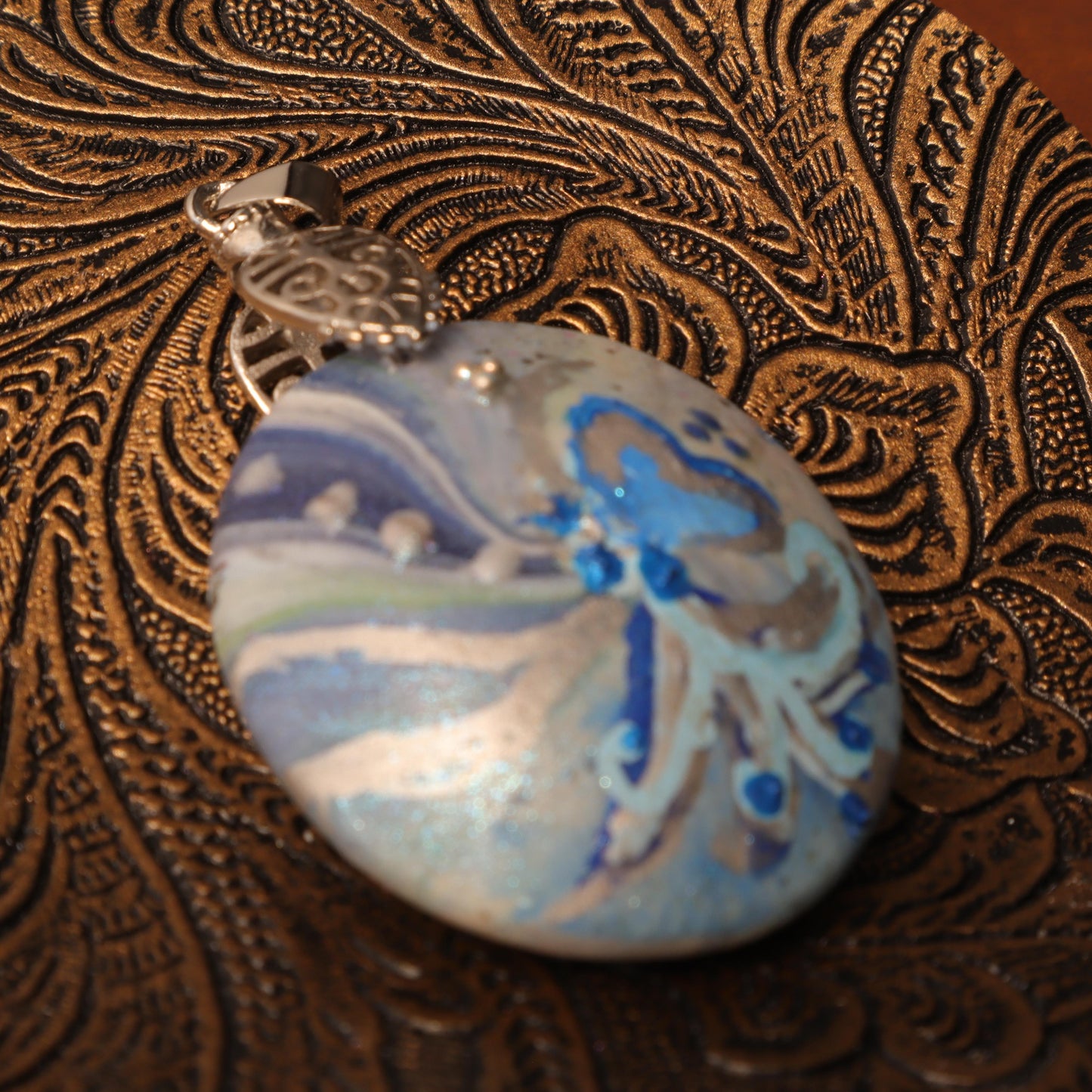 Close-up of a hand-painted polymer clay pendant with ocean-inspired swirls in blue and silver hues